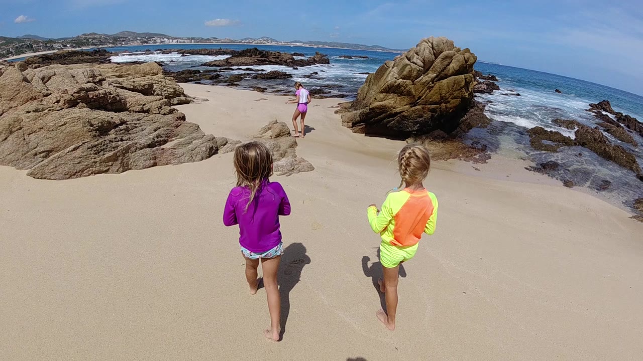At the start of the Cabo clip, two children seen from behind run over pale sand toward a large tan rock and white surf, with another child farther ahead.