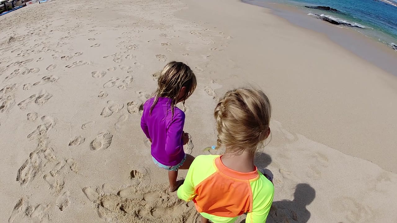 Two children are viewed from above on a Cabo beach, standing by a small uneven patch of sand while the ocean remains visible along the upper right.