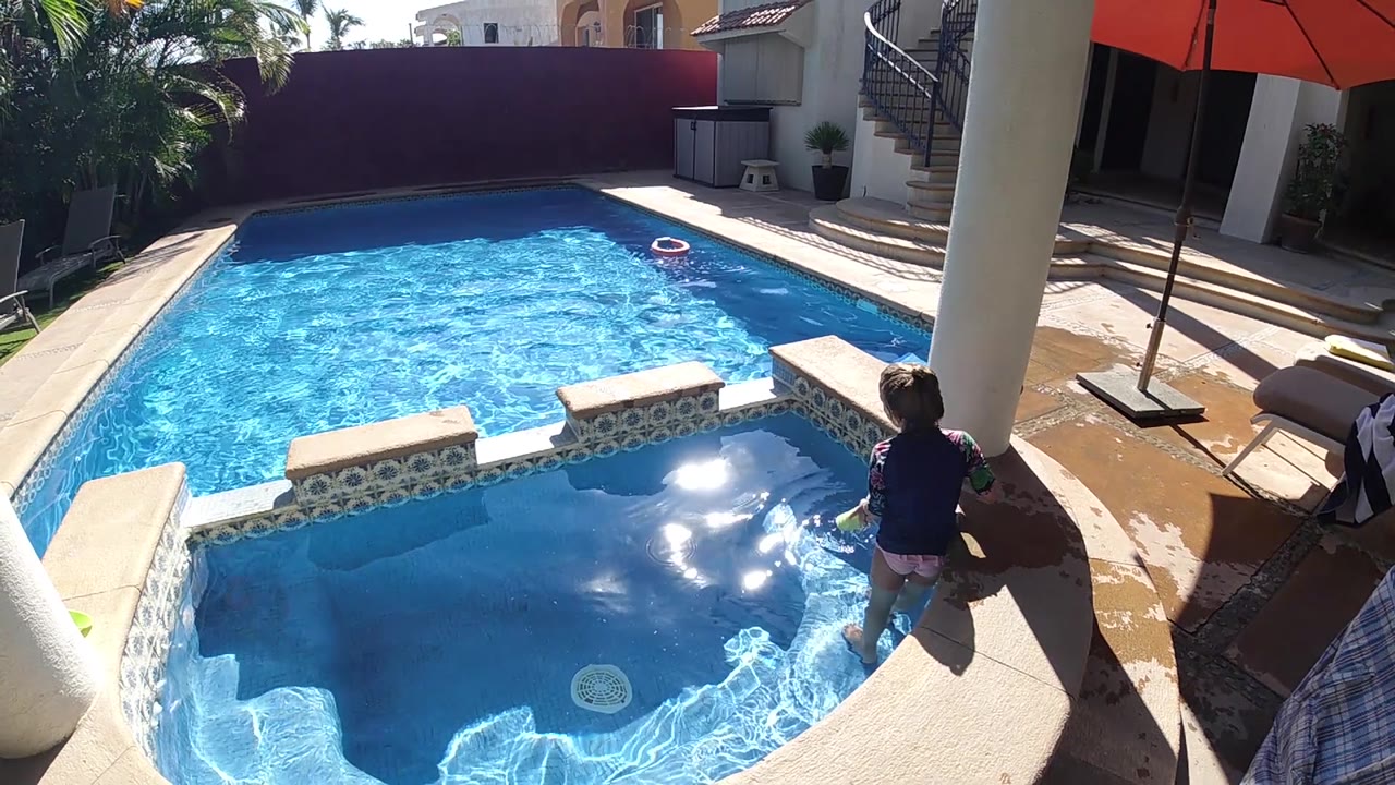 A child at the shallow pool edge faces the main pool area, with blue water, a floating ring in the distance, patio seating, and a red umbrella visible around the residential deck.