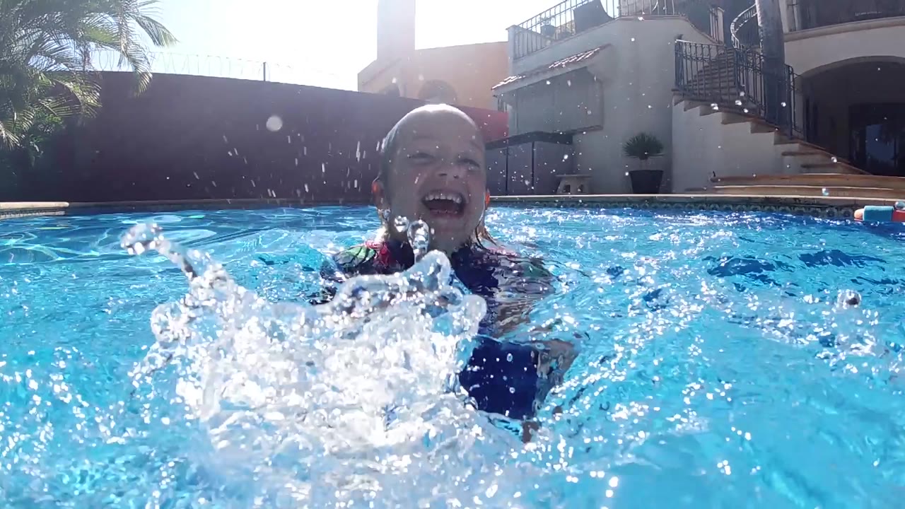 The boy laughs above the water as a lower burst of spray crosses his chest, with sunlit ripples and the poolside house behind him.