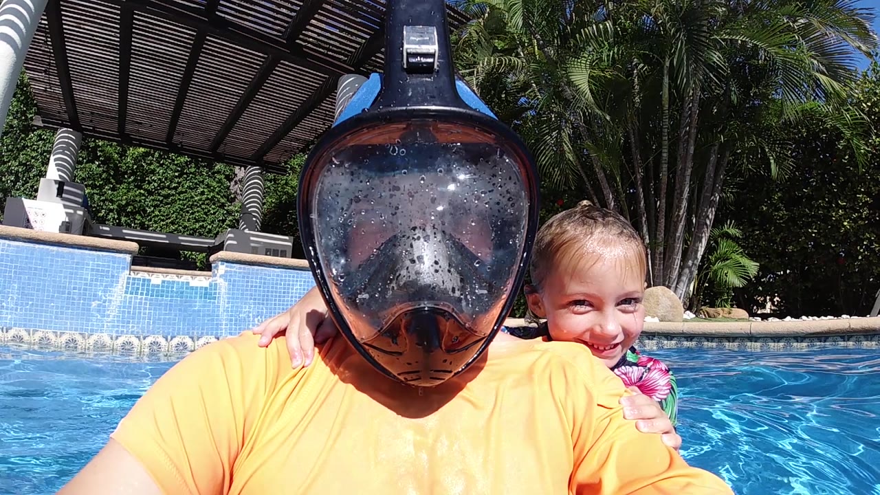 At 0:02, the adult's wet snorkel mask dominates the foreground while the child smiles from the right side of the frame in the sunlit Cabo swimming pool.