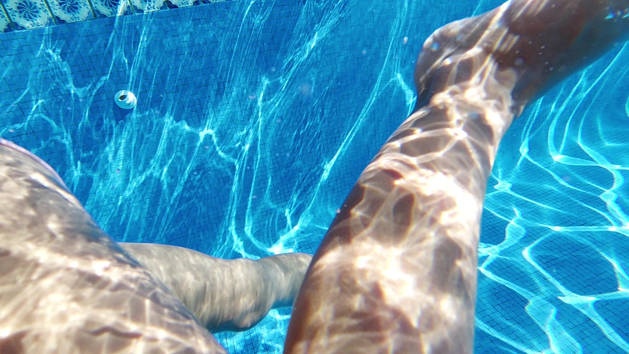 Underwater Cabo pool frame with two swimmer legs near the surface, bright caustic light on skin, blue tile border, and a small round pool fitting visible.