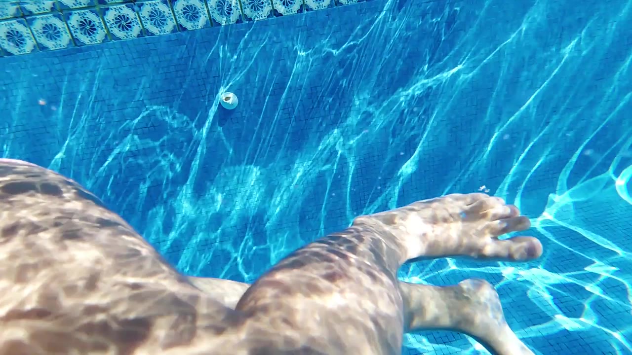 Underwater Cabo pool frame with a forearm and hand extending to the right, a leg crossing the left side, clear blue water, and rippled light on the pool wall and floor.