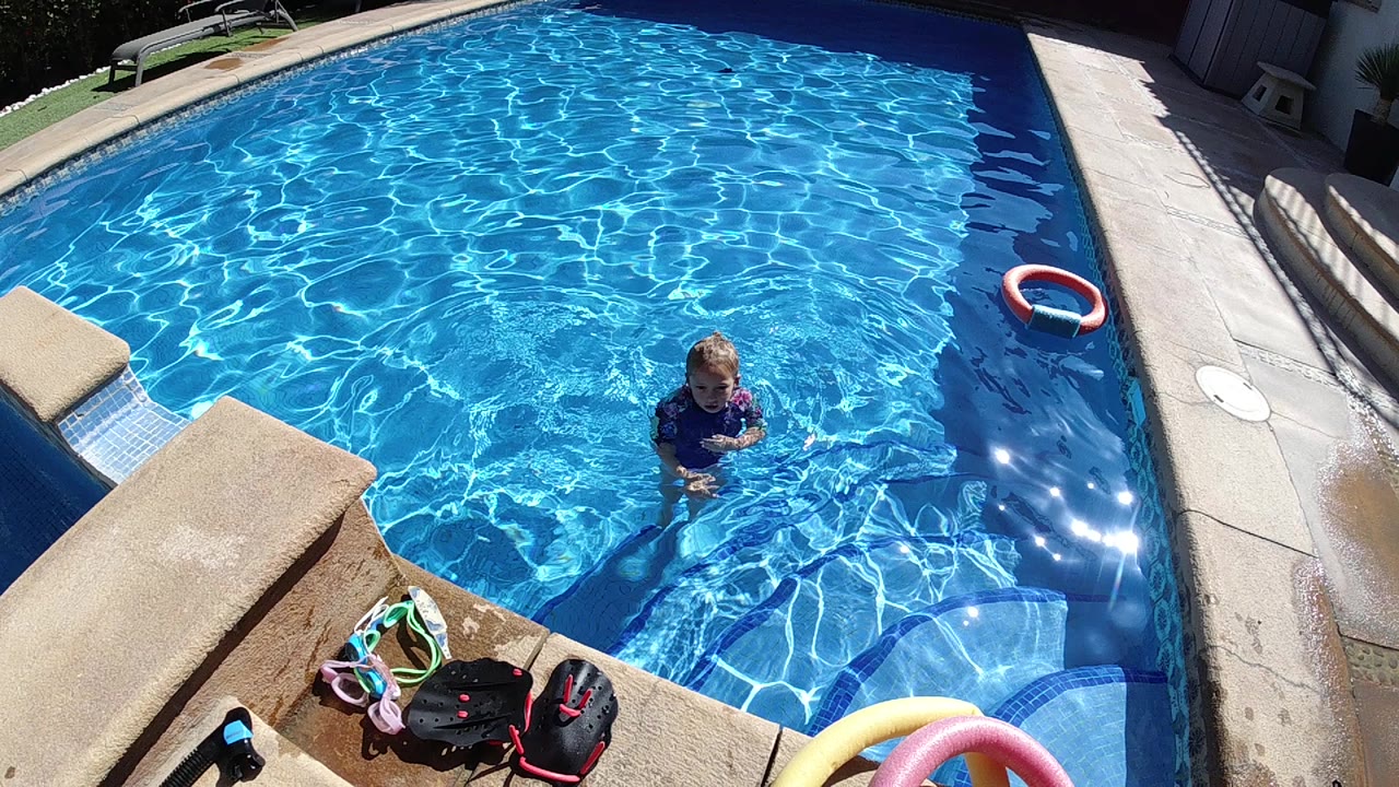 High-angle view in Cabo, Baja California Sur of a child in a dark swim shirt standing in clear blue pool water, with pool steps, deck coping, toys, and a red and white float visible.