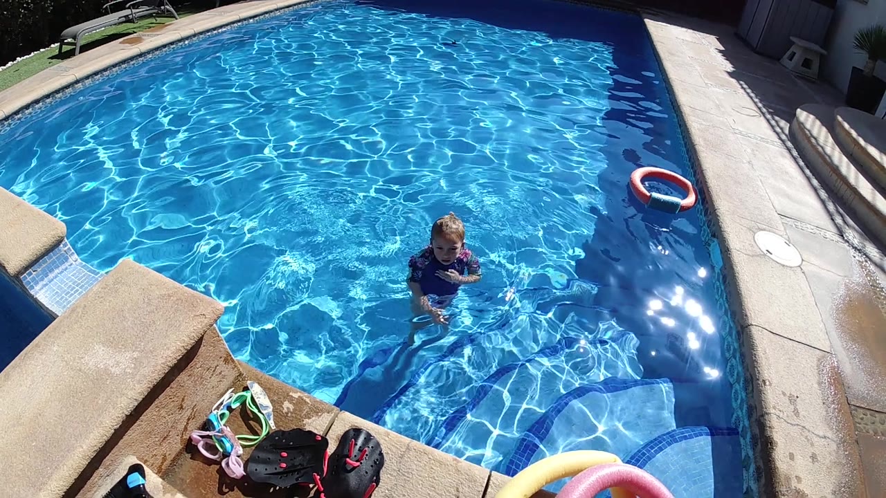 Cabo pool frame from above with the child in dark swimwear near the lower right, bright blue water across the frame, deck edge, pool toys, and a red and white float at the side.