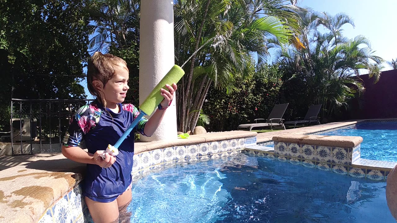 Young girl in a blue swimsuit holds a green water gun near the pool edge in Cabo, with bright water, patterned tile, palms, and a white pillar behind her.