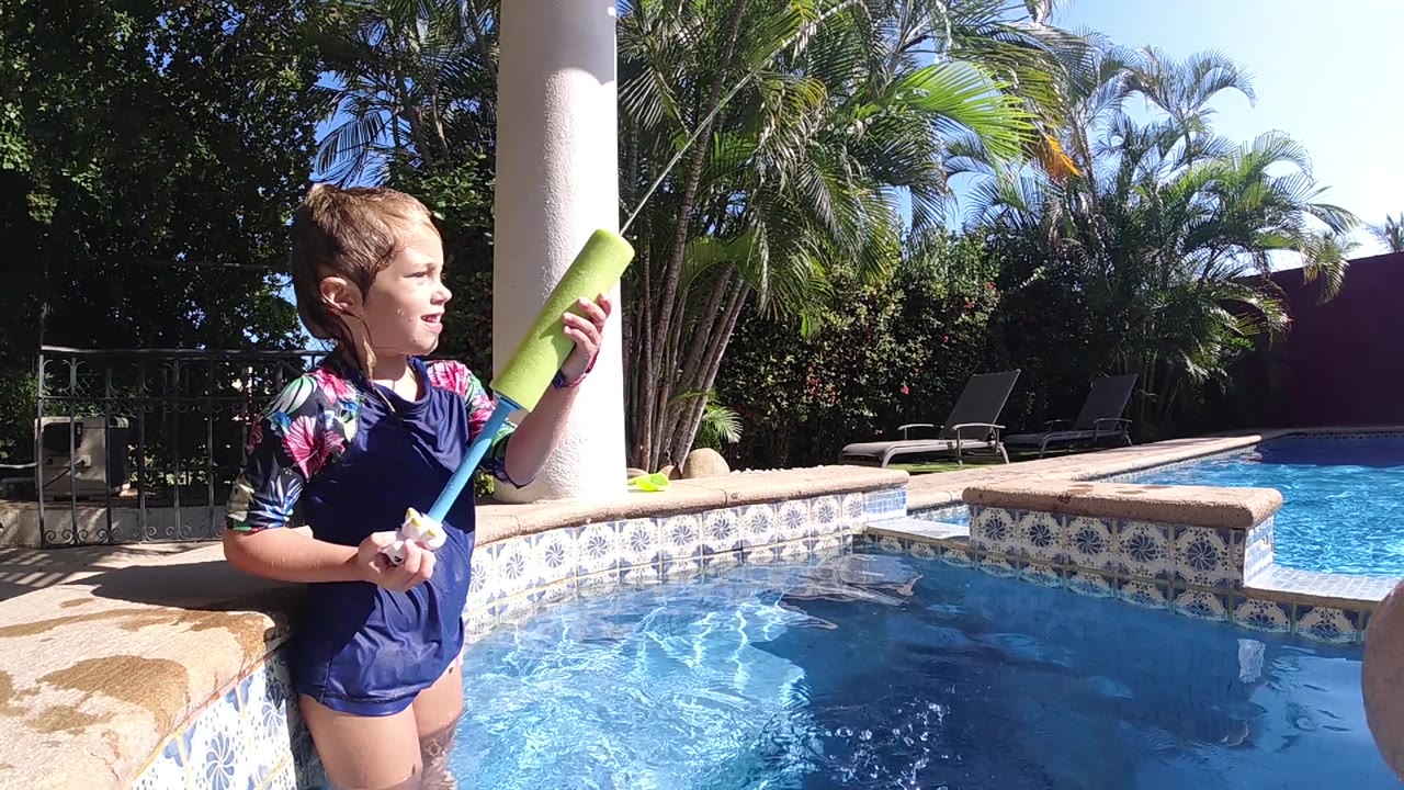 The girl turns her face toward the pool while holding the green water gun upright, with rippling blue water and palm fronds filling the Cabo patio scene.