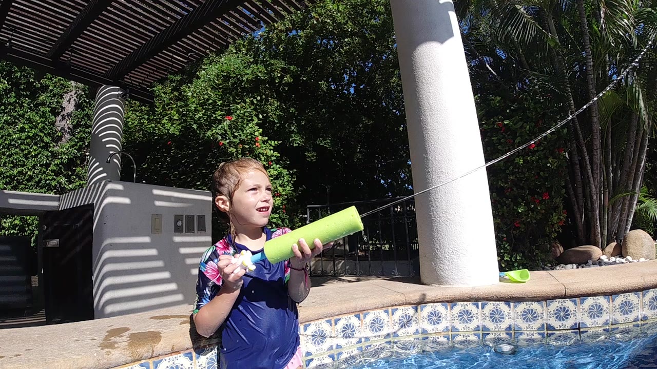 A young boy in a blue floral swim shirt holds a green pool noodle by a tiled pool in Cabo, Baja California Sur, with sunlit water and a white column behind him.