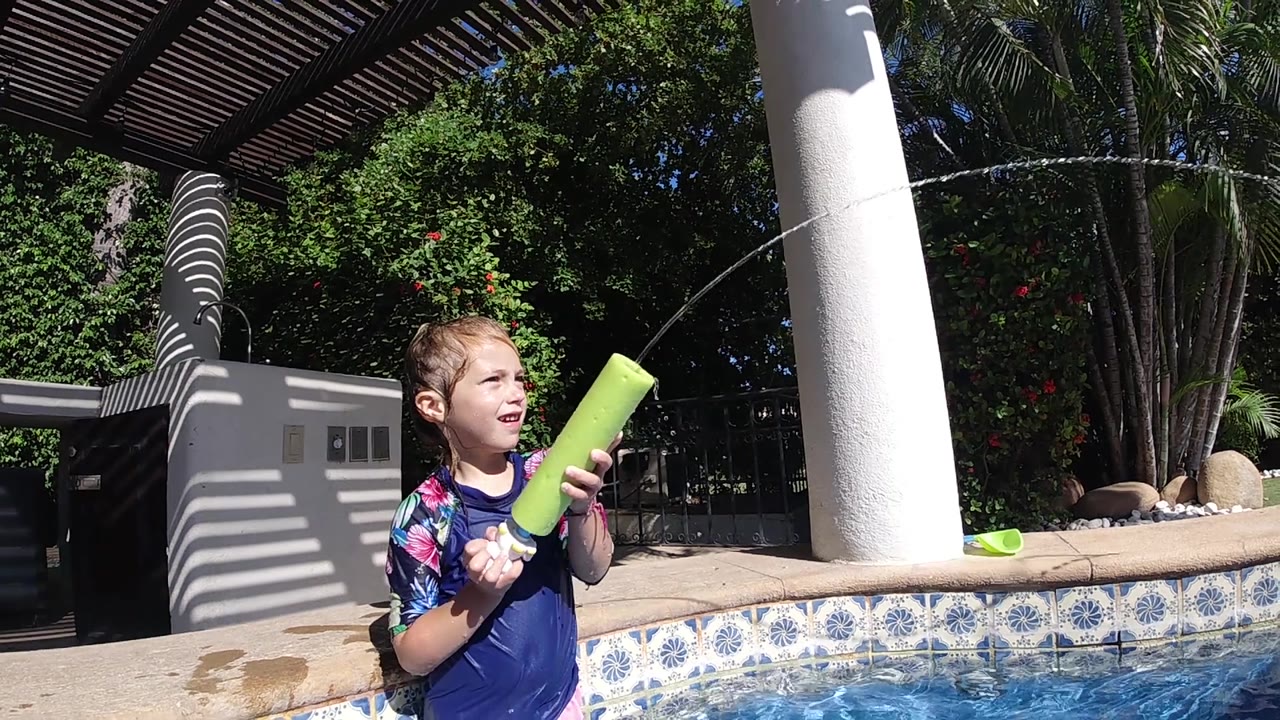 A child in a blue swim shirt grips a green pool noodle beside the pool in Cabo, Baja California Sur, with a dark pergola roof and leafy background behind him.