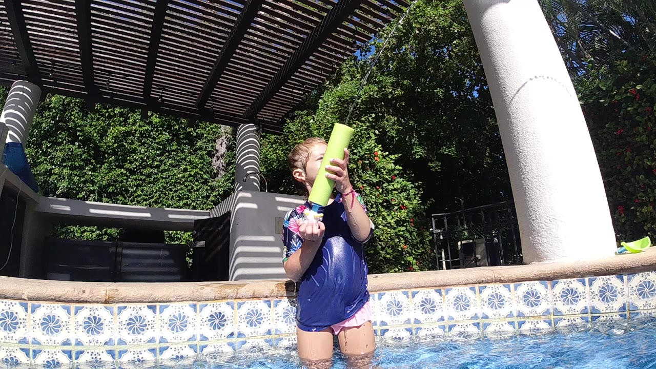 A young girl in Cabo, Baja California Sur stands waist-deep in a swimming pool and holds a yellow water gun upright near her face beneath a slatted pergola.