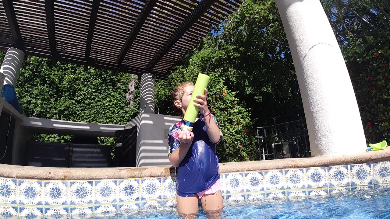 Girl in blue swim shirt holds a yellow water gun by the pool edge.