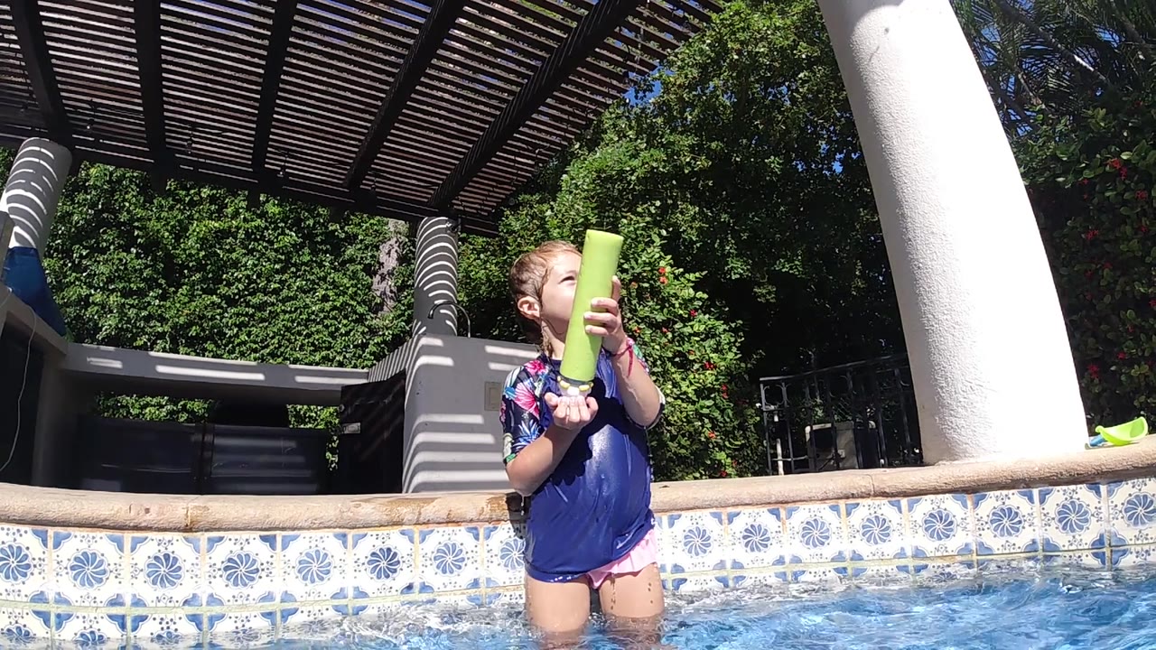 The girl looks upward with the yellow water gun angled near her shoulder, standing in bright pool water below the dark pergola in Cabo, Baja California Sur.