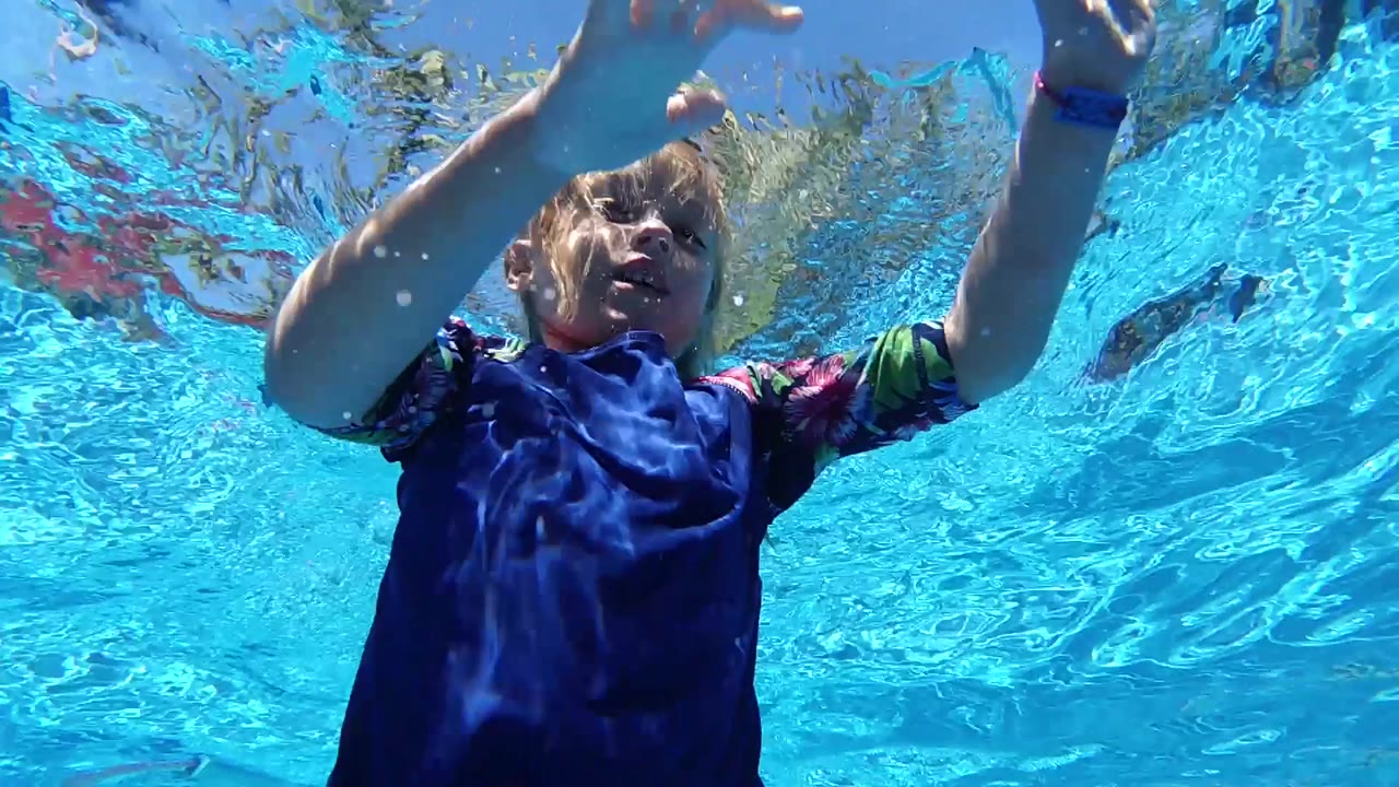 At 0:02, the child drifts under the pool surface with one hand high in frame and the face softened by moving water in Cabo, Baja California Sur.