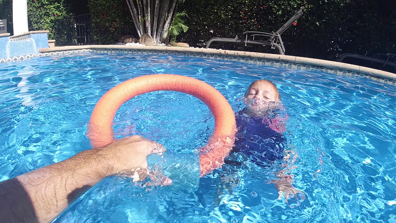 Adult hand holds an orange ring as the child floats nearby in a swimming pool.