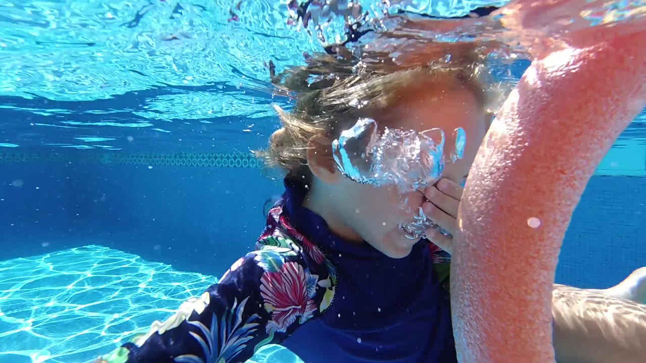 A close underwater view shows the child's face partly hidden by rising bubbles beside the orange noodle in the blue pool.