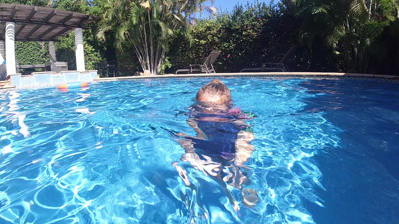 At the start, a child in a blue life vest faces away while floating in a bright blue pool in Cabo, Baja California Sur, with a patio and greenery beyond the water.