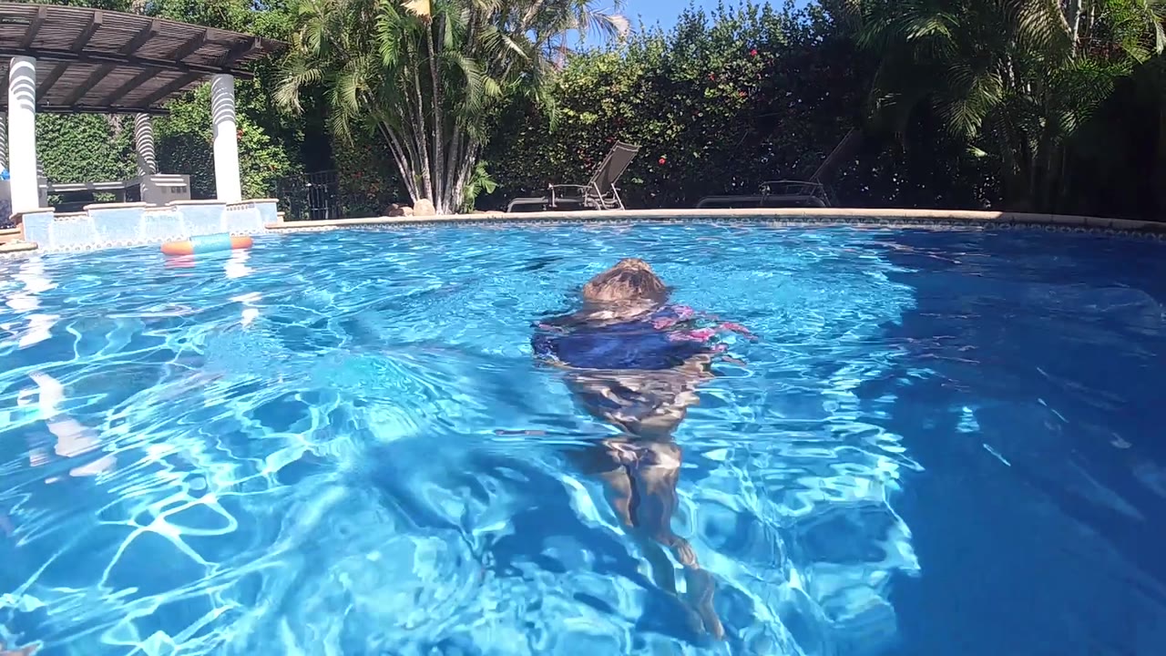 The child drifts farther from camera in the blue Cabo pool, with their head and vest visible above rippled water and shaded greenery behind.