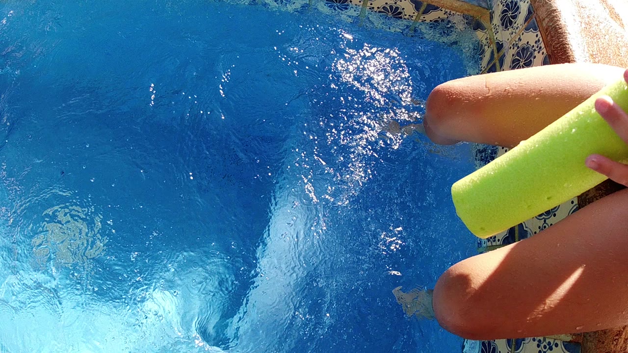 At the start of the Cabo poolside clip, bare legs and feet sit at the right edge above bright blue rippling water with a lime green cup partly visible.