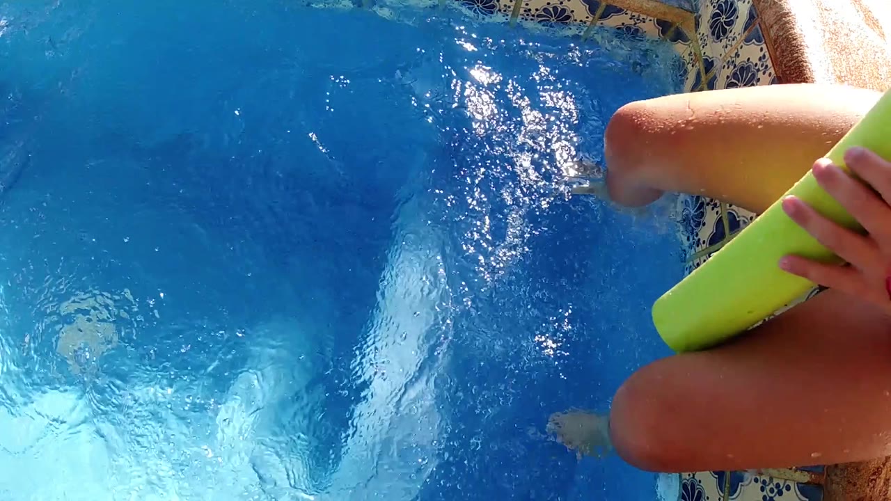 The cropped poolside figure in Cabo holds the green cup near bent legs while sunlight flashes across the pool surface.