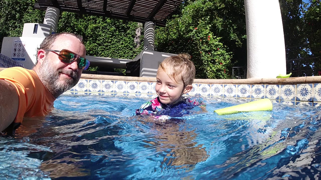 At the start of the Cabo clip, a man in sunglasses and an orange swim shirt floats at water level beside a young boy near a yellow pool noodle.