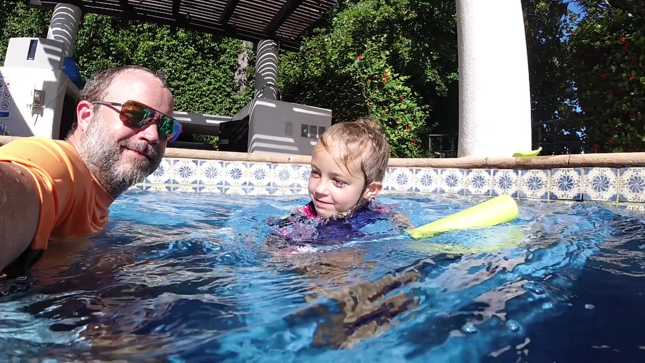 The boy turns toward the man as they float in clear water, with the yellow pool noodle close by and strong sunlight on the patterned pool wall.
