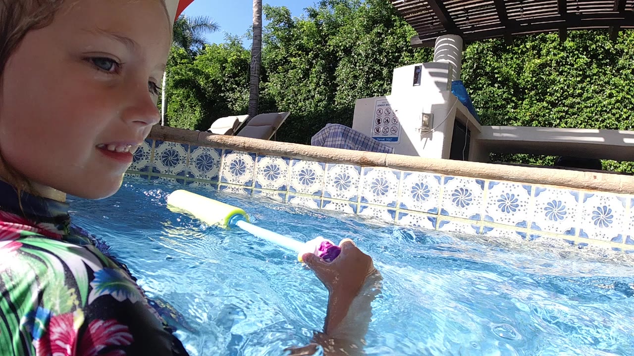 A young girl in a floral swimsuit holds a foam water toy near the blue pool surface in Cabo, Baja California Sur, with patterned tile and patio furniture behind her.