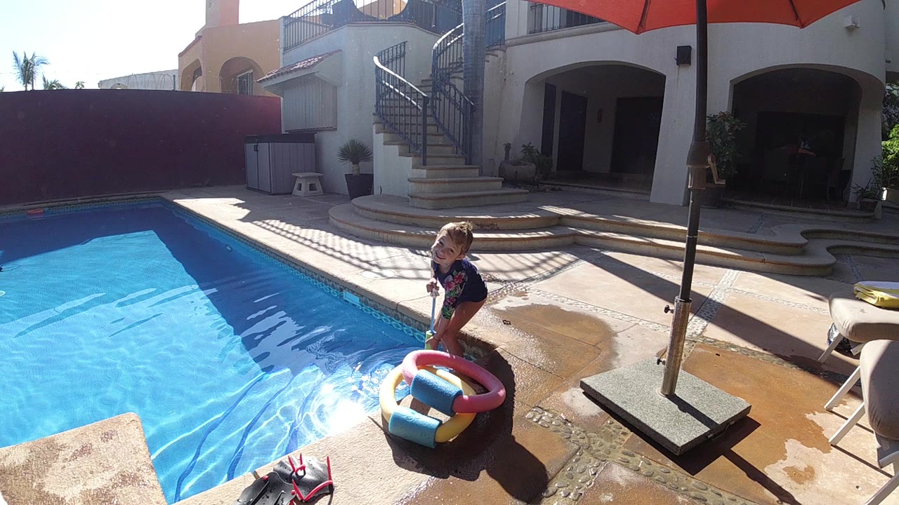 In Cabo, Baja California Sur, a child stands on a wet pool deck beside a colorful inflatable ring with bright blue water to the left and a red umbrella above.