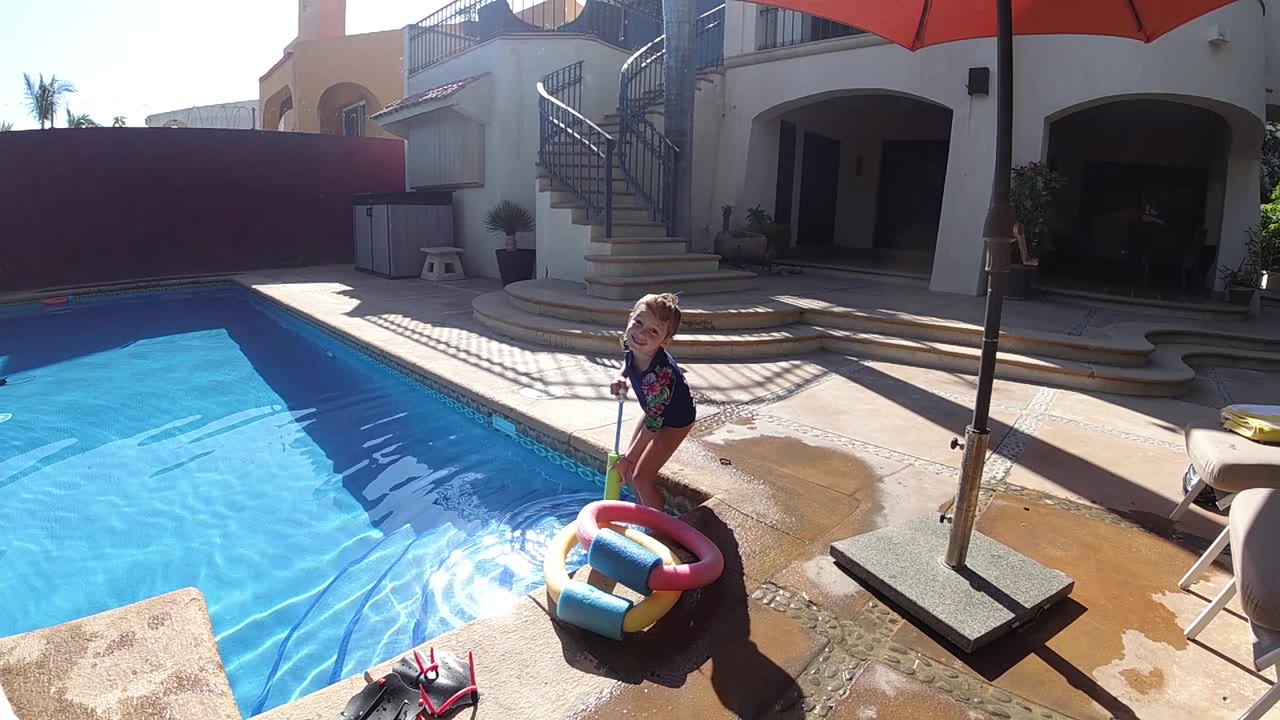 A wide poolside view shows the child upright near the colorful inflatable ring, with hard sunlight, wet concrete, and a shaded residential patio behind.