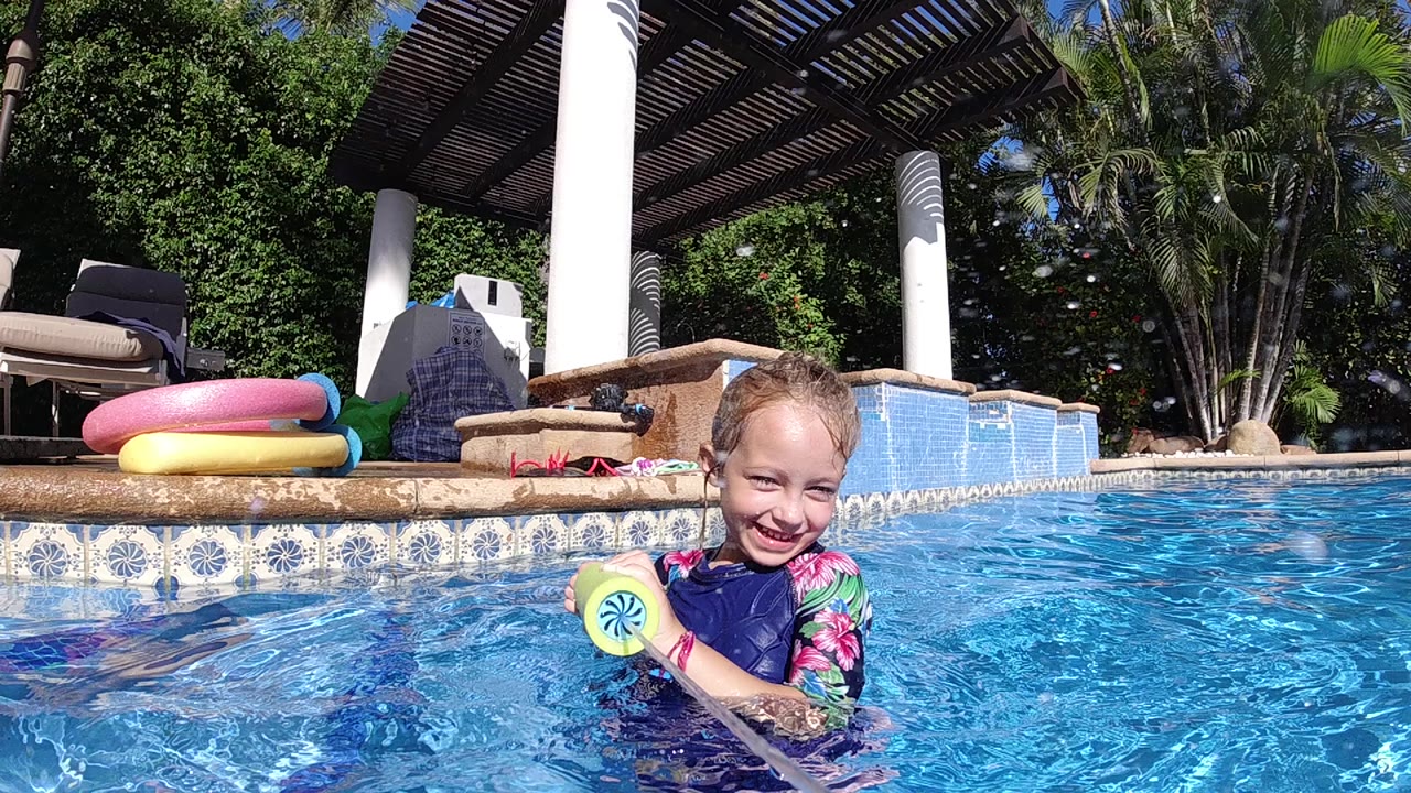 A toddler girl smiles at water level in a blue swimming pool in Cabo, Baja California Sur, holding a yellow toy with lounge chairs and inflatable rings behind her.