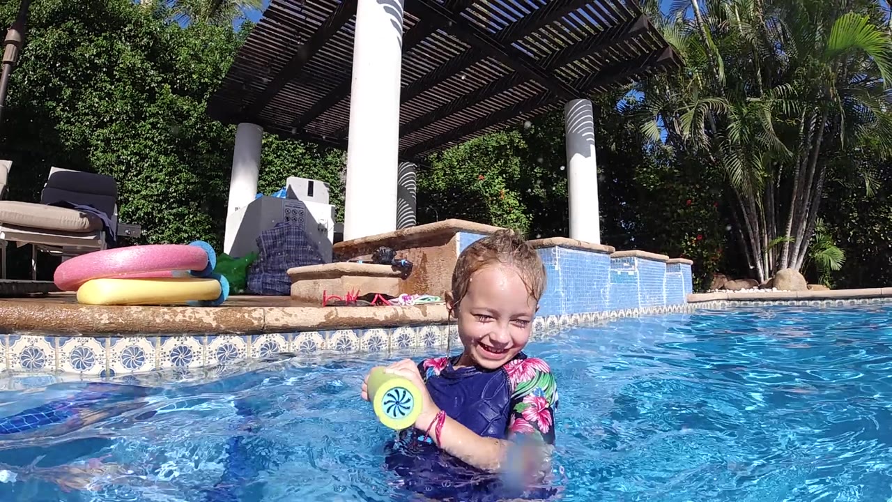 A toddler in a blue floral swimsuit stands in the pool in Cabo, Baja California Sur, with the yellow toy at chest height and the tiled pool edge behind her.