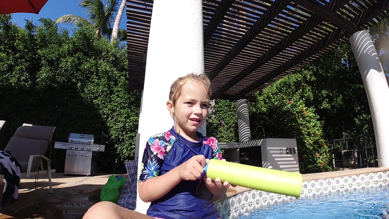A young girl in a floral-sleeved swim shirt holds the yellow water gun across her lap as the Cabo pool water catches bright sun at frame right.