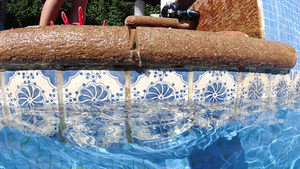 Wooden log water feature at a Cabo resort pool, decorative blue and white floral tiles lining the pool wall, blue water in lower frame; a red object at the upper edge may be footwear on the nearby deck.
