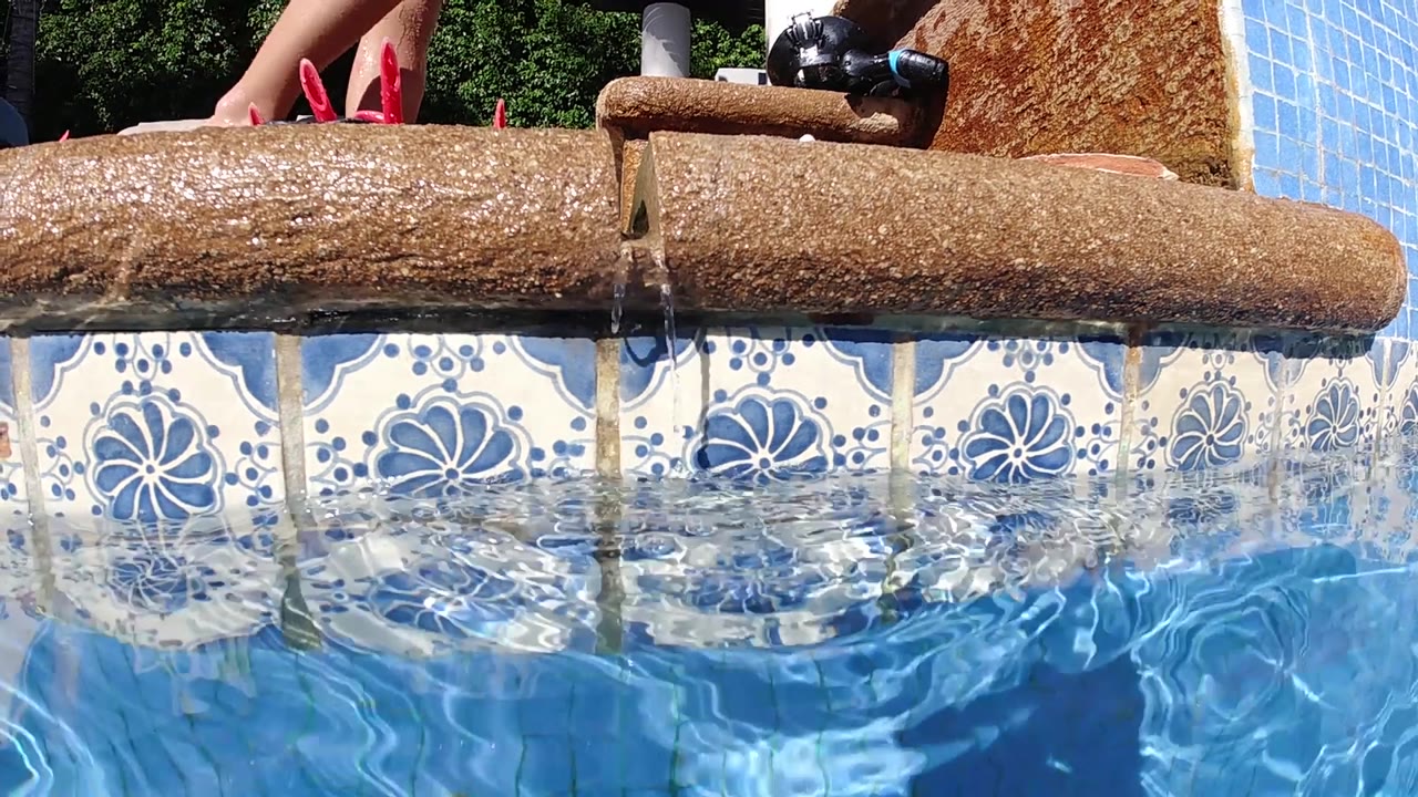 Rustic log positioned above white tiles with blue floral medallion patterns at a Cabo pool; bright blue rippling water fills the lower half and a red sandal or object is visible at the upper frame edge.