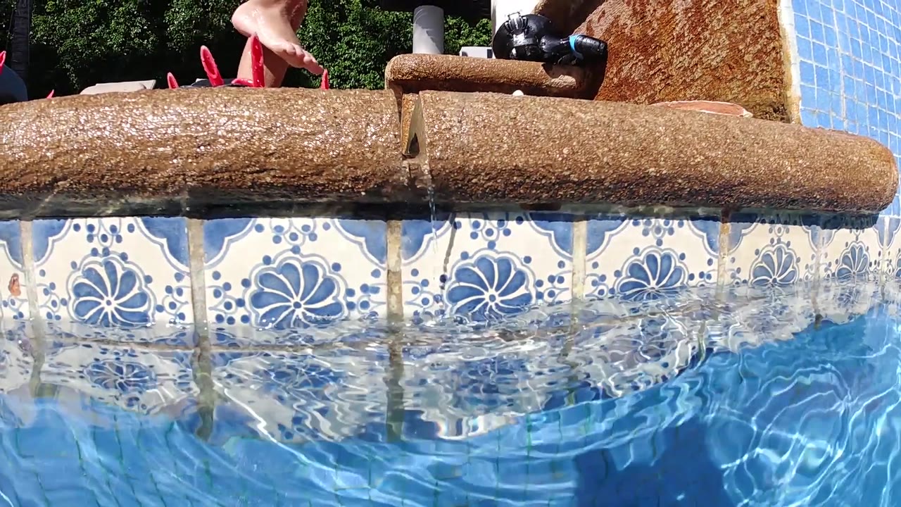 Rustic log at pool edge, decorative blue and white tile wall below, bright blue water, red footwear visible at top.