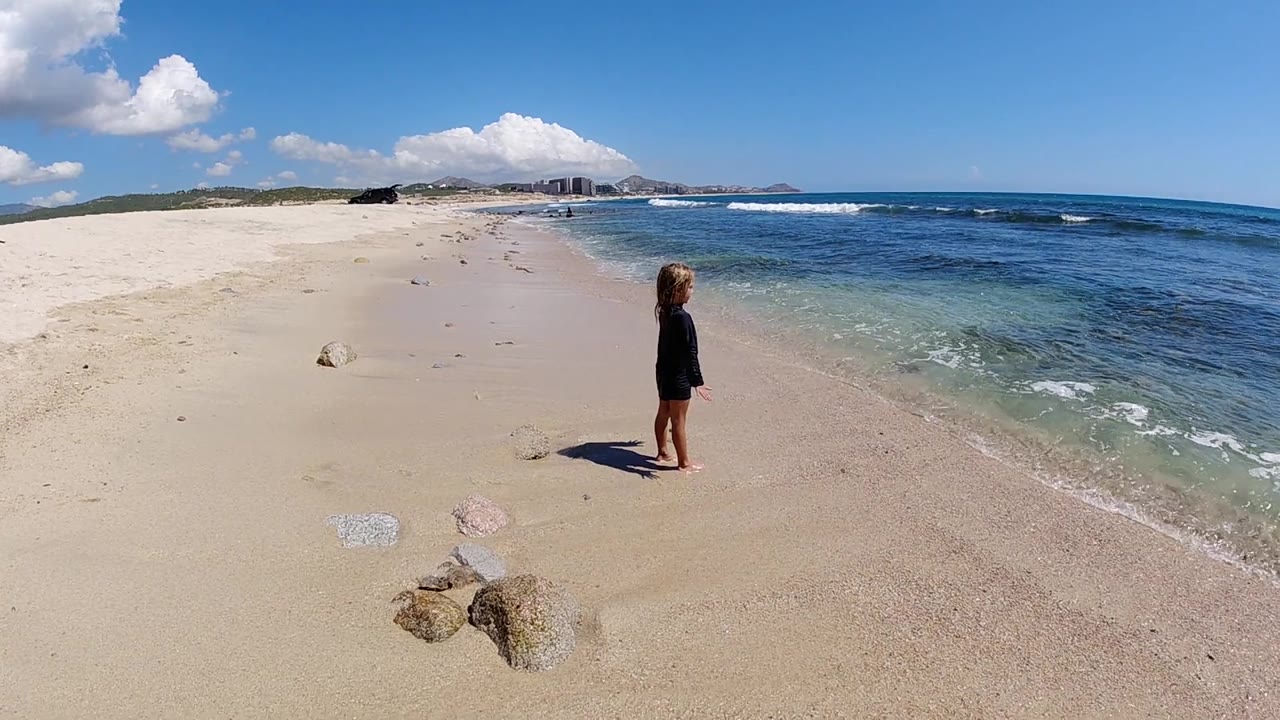 A child faces the blue ocean from the wet sand, with a thin wash of surf approaching and open beach stretching into the distance in Cabo.