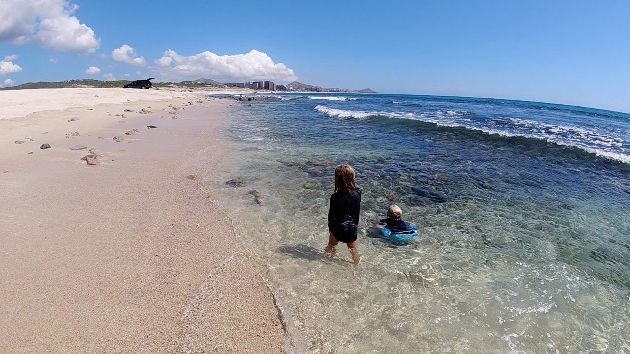 Two children in dark swim tops play in clear shallow surf on a Cabo beach, with pale sand to the left and blue ocean to the right.