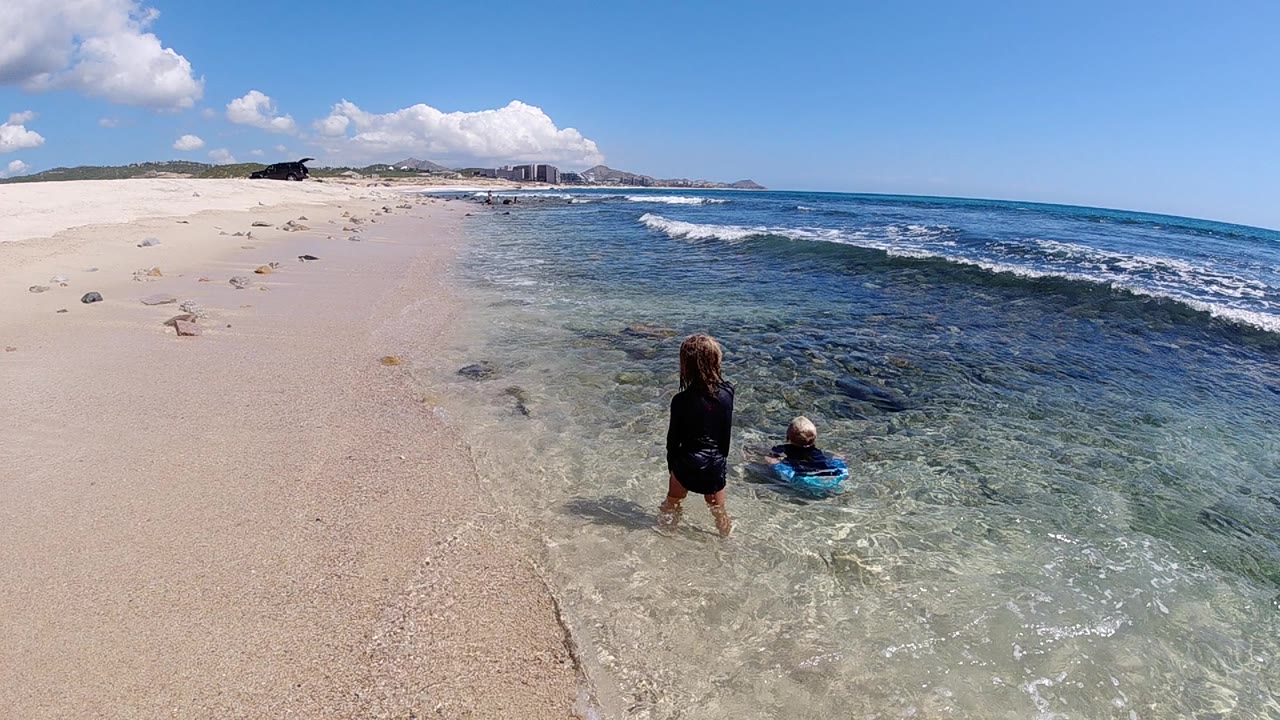Standing child watches a seated child in shallow beach water.
