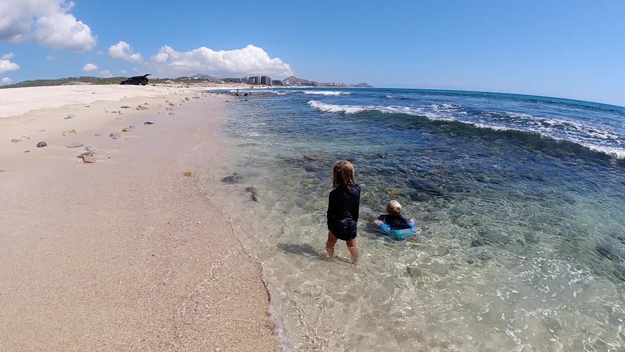 The two children stay near the water's edge in Cabo, with transparent shallows over sand and darker reef patches offshore.
