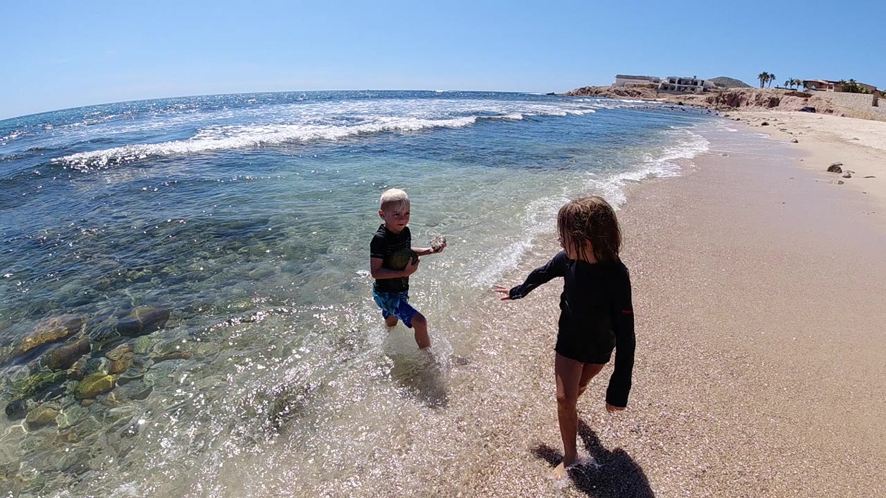 In Cabo, Baja California Sur, two children face each other at the edge of clear shallow water as small waves wash over the sand.
