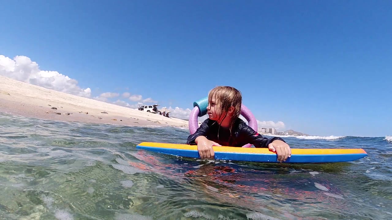 At the start of the Cabo clip, a young girl in a black wetsuit rests on a blue and yellow bodyboard while looking left across shallow water.
