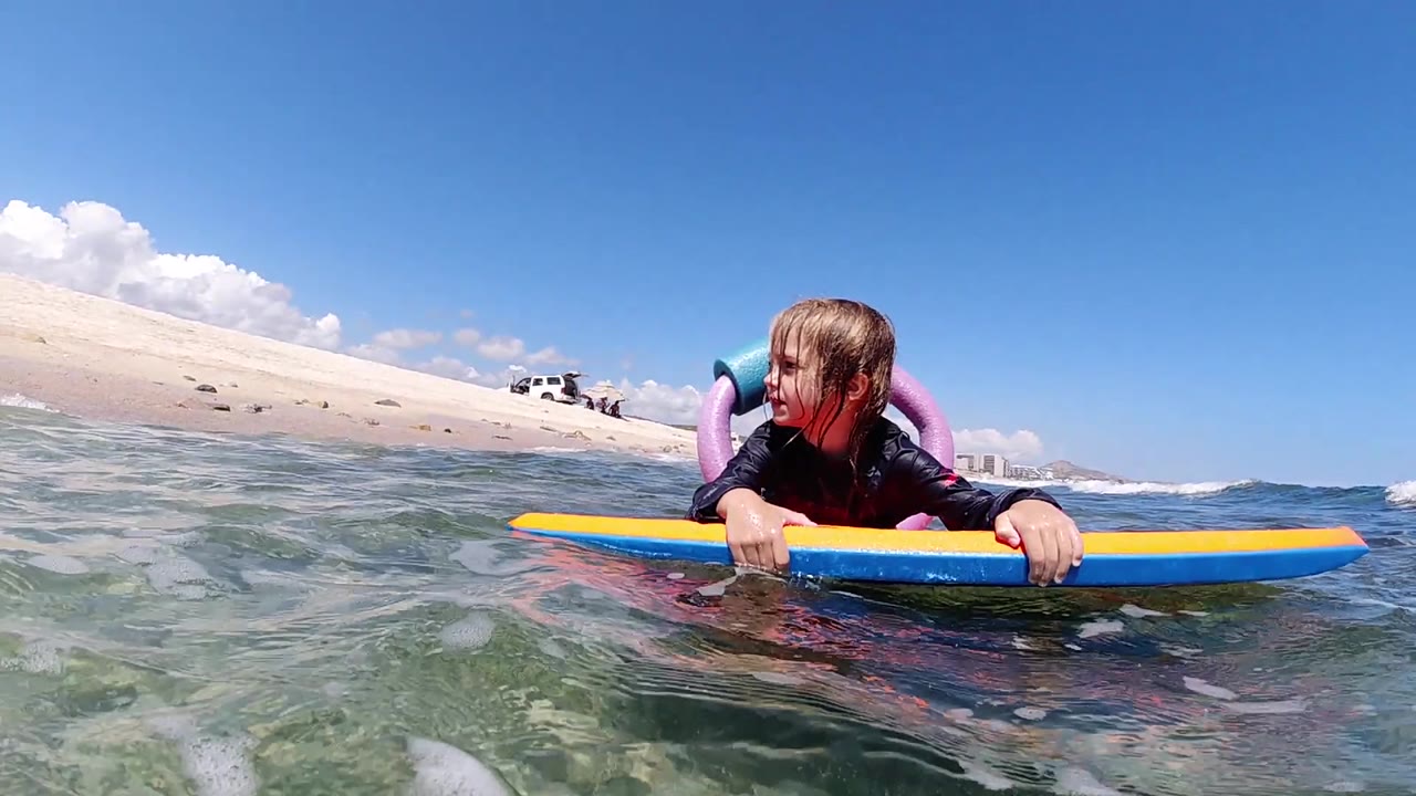 The child remains low on the board as the blue and yellow nose cuts across the shallow green water with pale sand rising behind her.