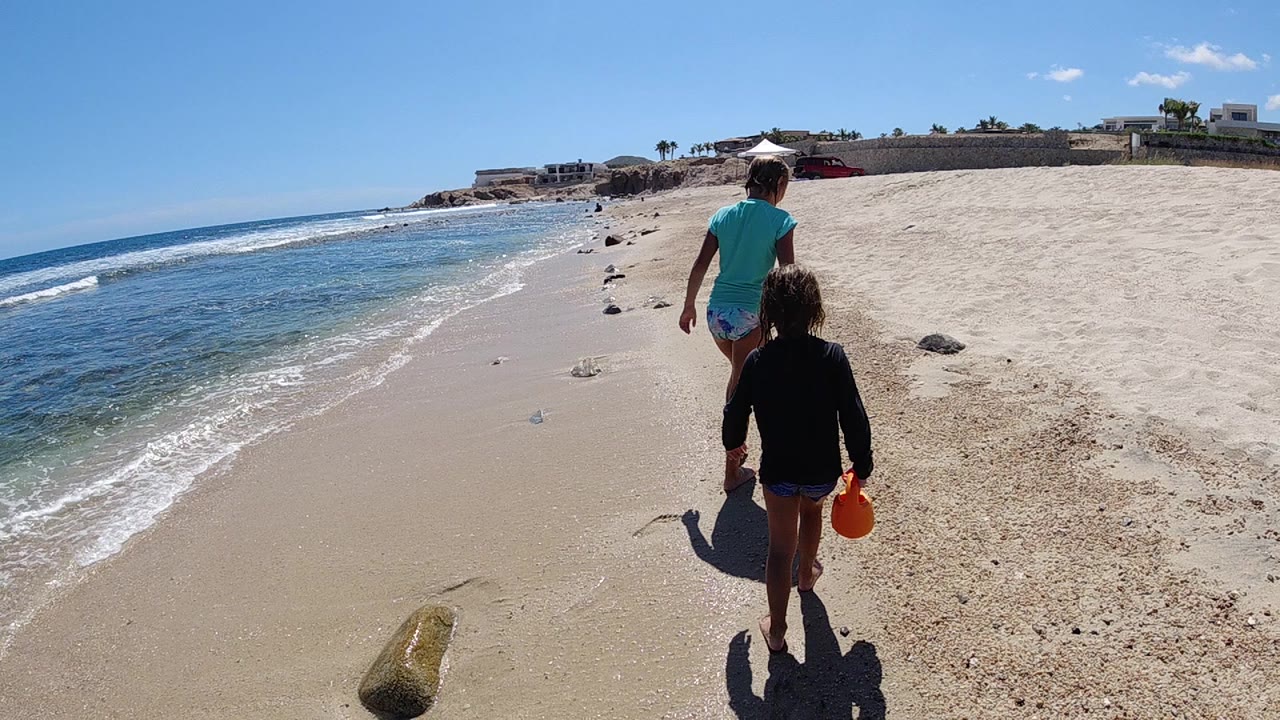 In Cabo, Baja California Sur, a man and child walk away from the camera near the shoreline, with the child holding a red bucket and a small log or rock in the lower sand.