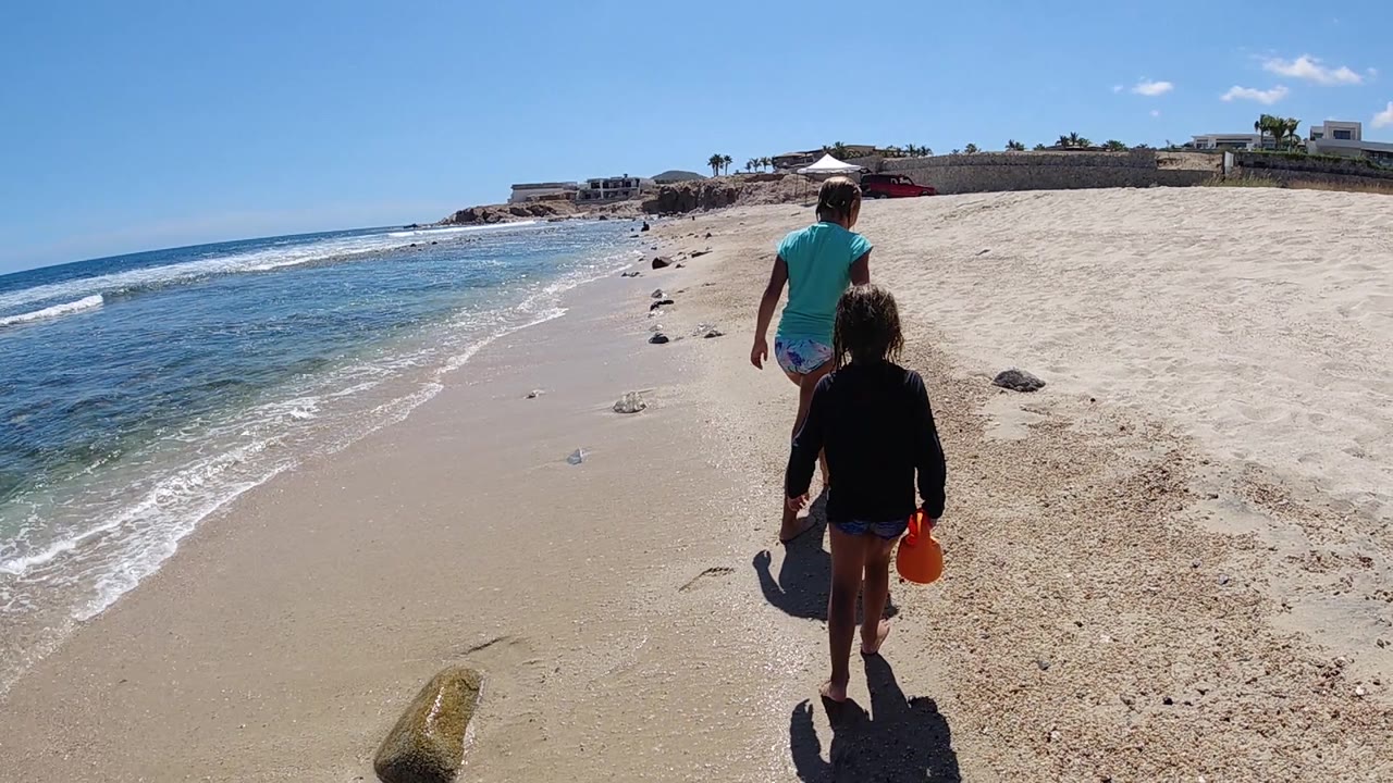 A child carrying a red bucket follows a man in a turquoise shirt along the Cabo sand, with the ocean to their left and low buildings far ahead.