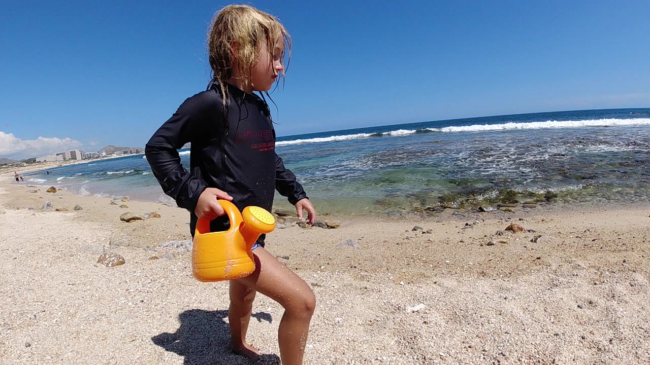 In Cabo, Baja California Sur, a child in a black rash guard walks across pale sand carrying a yellow watering can, with shallow ocean water and surf behind them.
