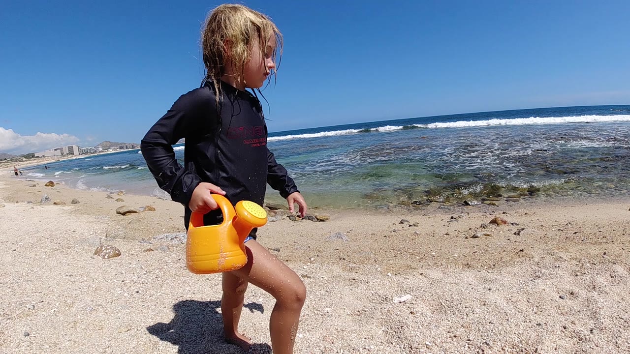 The child continues walking along the sandy Cabo beach, holding the bright yellow watering can low beside one leg as white surf crosses the background.