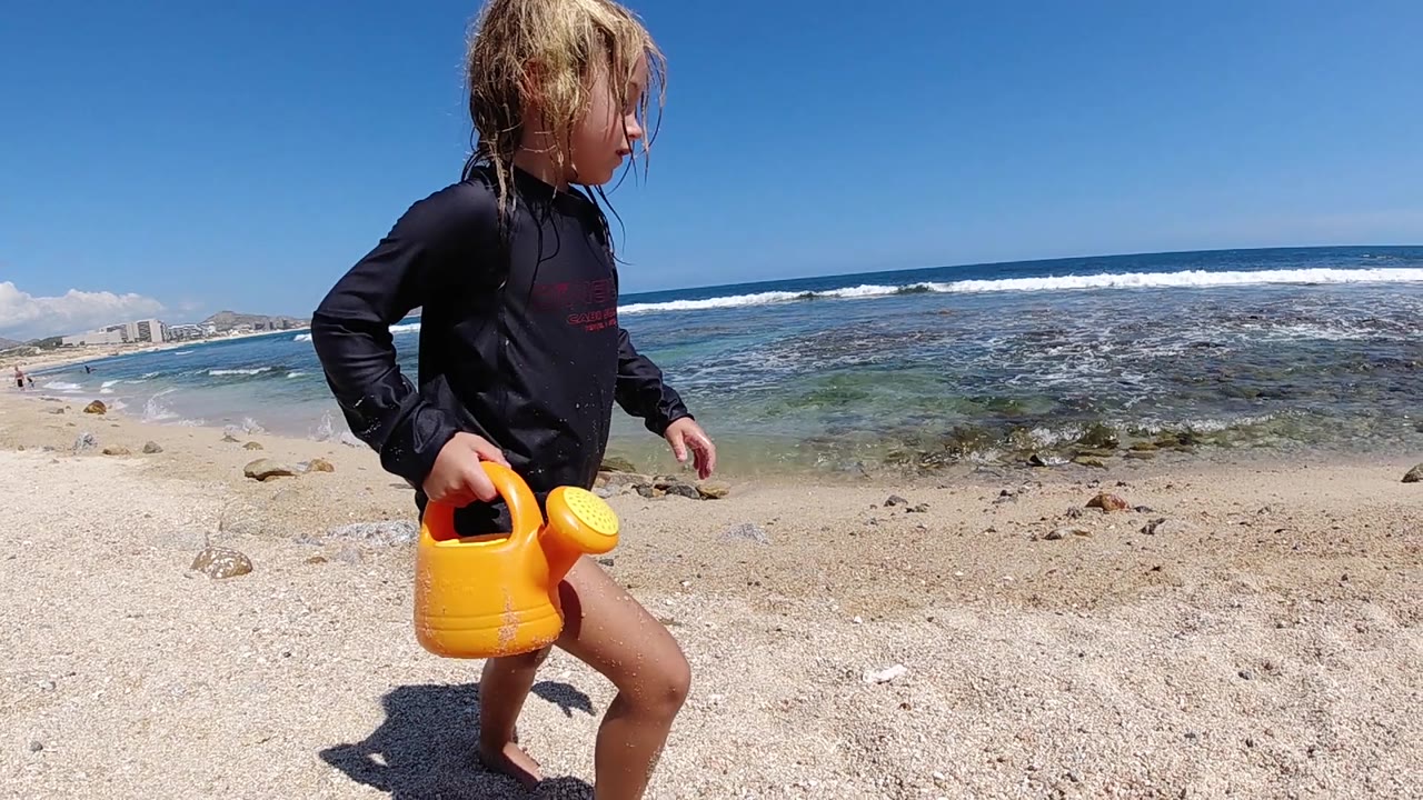 The child remains foreground right on the Cabo sand with wet hair, black rash guard, blue shorts, and the yellow watering can, while low waves break offshore.