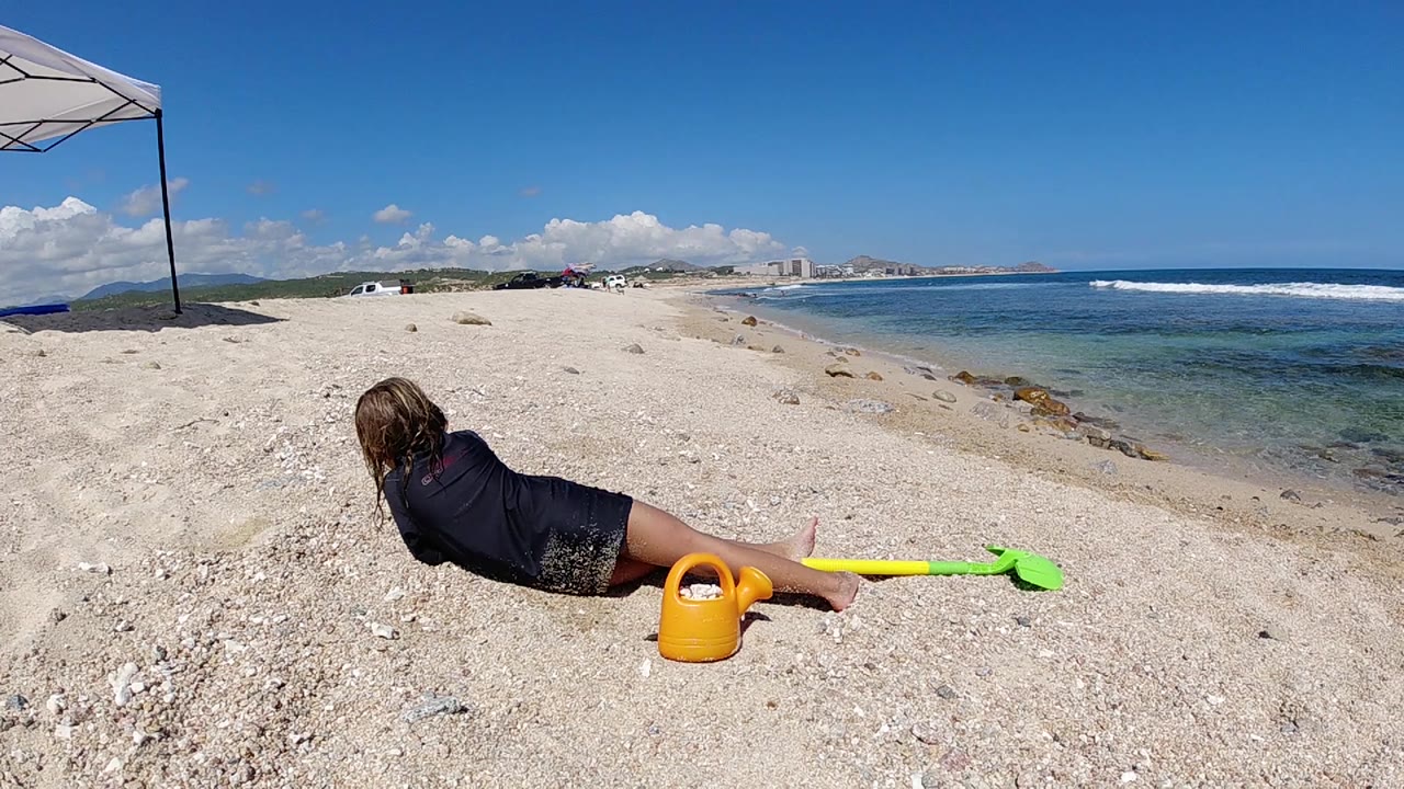 Child lies on their stomach on a Cabo beach beside a yellow watering can and green shovel, with blue water and distant shoreline behind.