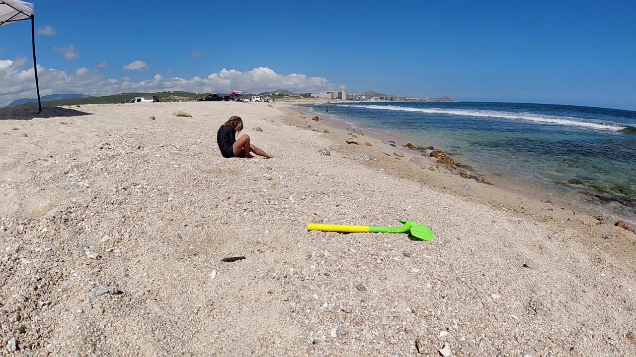 At the start of the Cabo beach clip, a seated person faces the water while a bright yellow and green toy shovel lies on pale sand in the foreground.
