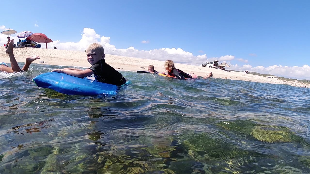 Child holds a blue boogie board while other children float behind.