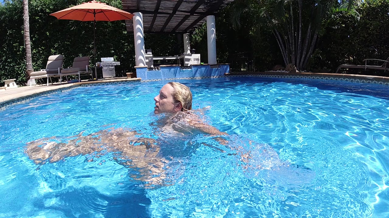 In Cabo, Baja California Sur, a blonde woman swims left to right through bright blue pool water with lounge chairs, a red umbrella, and greenery behind her.